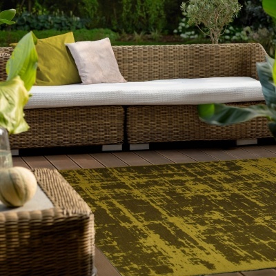 Sofá de vime castanho com almofadas verdes e bege, tapete verde e mesa pequena em ambiente exterior