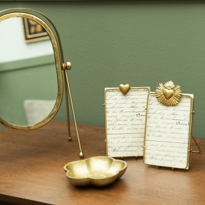 Espelho oval dourado, dois porta-fotos dourados com fundo de papel manuscrito e suporte decorativo dourado em superfície de madeira.