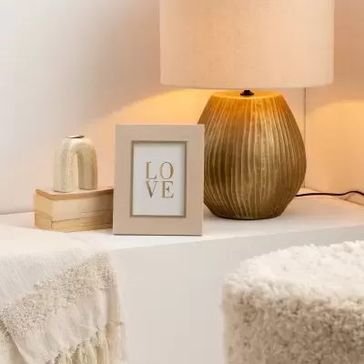 Lâmpada de mesa dourada com candeeiro bege, porta-retratos com texto LOVE, livros e peça decorativa numa mesa branca com tecido e puff peludo.