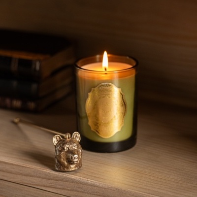 Vela perfumada acesa em copo de vidro verde com rótulo dourado e cabeça de urso dourada ao lado em madeira clara
