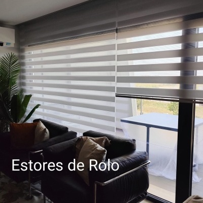 Estores de rolo brancos translúcidos numa sala com poltronas pretas e almofadas coloridas