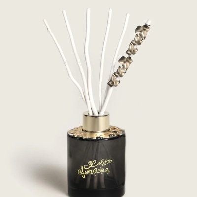 Difusor de ambiente em frasco preto com tampa dourada e pauzinhos brancos com um laço, texto Lolita Lempicka