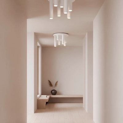 Corredor com chão de madeira clara, paredes brancas, candeeiros de tecto cilindricos brancos, banco em madeira clara, vaso negro com ramos secos e livro branco.