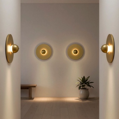 Arandelas douradas redondas iluminadas num corredor com chão de madeira clara, planta e banco de madeira