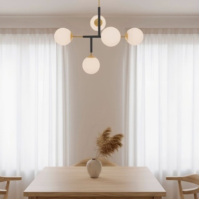 Luminária de teto com globos de vidro branco sobre mesa de madeira clara com cadeira e vaso com plantas secas.