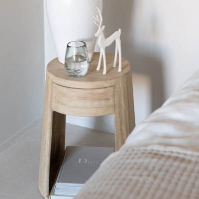 Mesa de apoio redonda em madeira clara com vaso branco, estatueta de rena e copo de água, perto de cama com cobertor bege