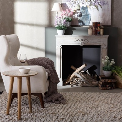 Cadeirão branco com manta castanha junto a mesa de madeira, lareira branca decorativa com lenha e plantas.