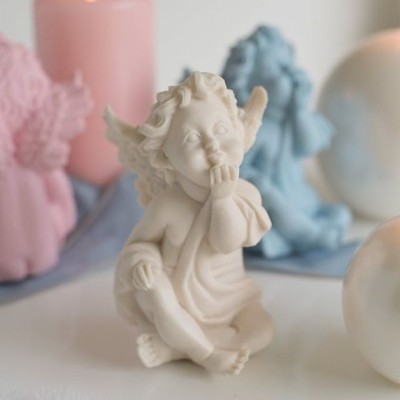 Figura decorativa de anjo branco sentado com mão no rosto, com vela rosa acesa e anjos coloridos ao fundo.