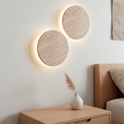 Apliques redondos de pedra bege com luz suave atrás, acima de mesa de cabeceira de madeira clara com vaso branco e ramo seco.