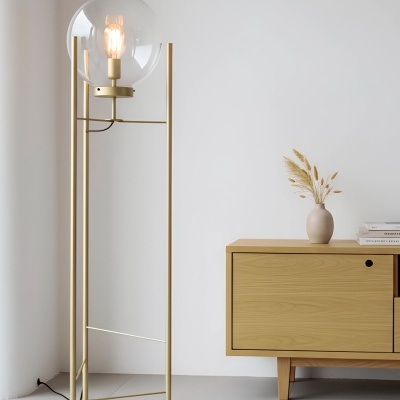 Candeeiro de pé dourado com cúpula de vidro ao lado de móvel baixo de madeira com vaso e flores secas