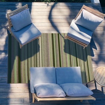 Conjunto de cadeiras e sofá de madeira clara com almofadas cinza claro sobre tapete verde às riscas numa plataforma de madeira ao ar livre