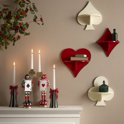 Prateleiras de parede em forma de espadas, paus, copas e ouros em creme e vermelho numa parede bege acima de lareira com velas e decoração