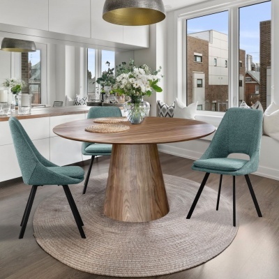 Mesa de madeira redonda com três cadeiras azuis e jarra de flores numa sala iluminada