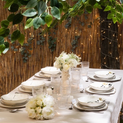 Mesa posta com pratos de cerâmica com padrão de folhas, copos de vidro e flores brancas numa mesa com toalha branca, decorada com luzes e parede de bambu