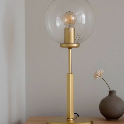 Candeeiro de mesa dourado com abajur de vidro esférico ao lado de vaso cinzento com flor