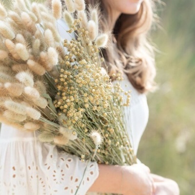 Mulher de vestido branco segurando ramo grande de flores secas em ambiente natural