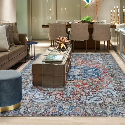 Sala de estar e jantar com sofá castanho, mesa de centro em couro, tapete floral azul e vermelho, cadeiras bege e candeeiros de velas