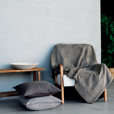 Poltrona de madeira com mantas e almofadas cinzentas ao lado de banco de madeira com prato branco em ambiente minimalista