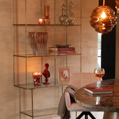 Estante metálica dourada decorada com vasos, esculturas, livros e velas em sala com cadeira e mesa.