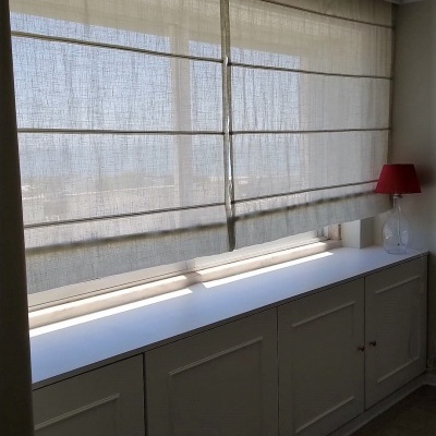 Janela com estores romanos bege, móvel branco com armários e abajur vermelho