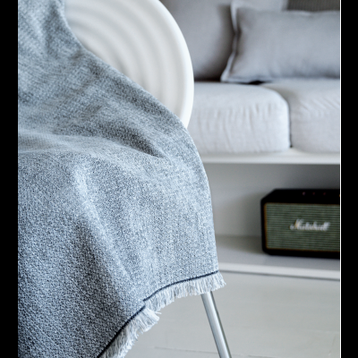 Cobertor cinzento com franjas numa cadeira branca num ambiente interior com sofá cinzento e coluna com coluna de som Marshall