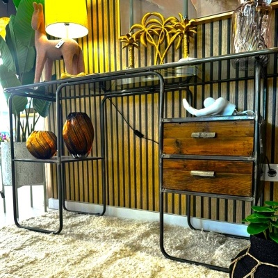 Mesa metálica com tampo de vidro, gavetas de madeira, abajur amarelo e objetos decorativos num ambiente interior