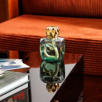 Difusor de perfume transparente com tampa dourada numa mesa de vidro com sofá laranja ao fundo