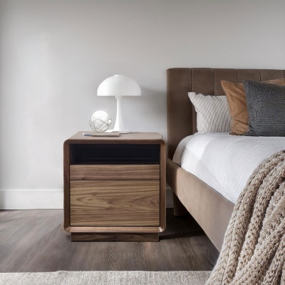 Mesa de cabeceira em madeira com candeeiro e decoração ao lado de cama estofada com almofadas e manta.
