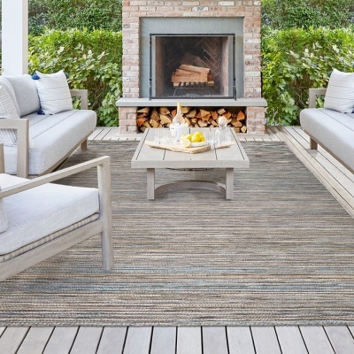 Zona de estar exterior com tapete cinza e bege, sofás, cadeira de madeira, mesa e lareira em tijolo