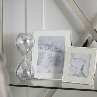 Quadros em moldura branca com imagens cinzentas e ampulheta prateada sobre superfície de vidro