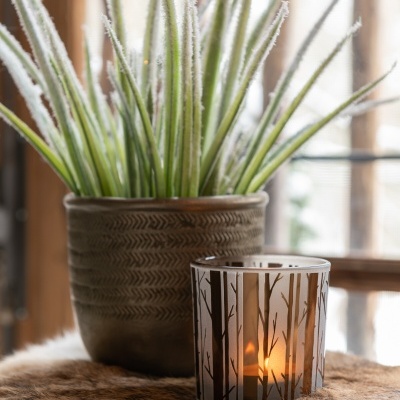 Vela acesa em jarra de vidro decorativa ao lado de vaso com planta sobre pele sintética