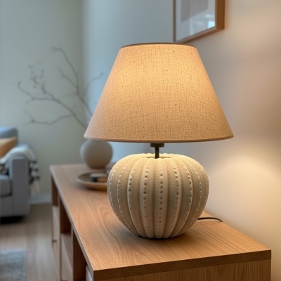 Lâmpada de mesa bege com base esférica e aba de tecido sobre móvel de madeira clara