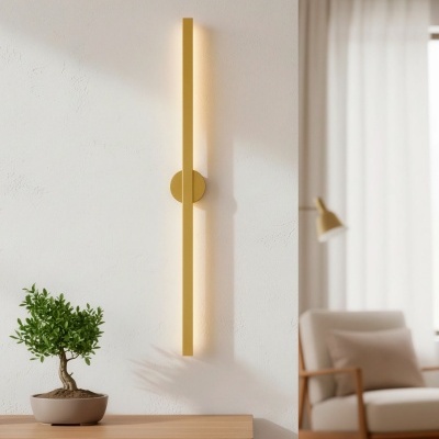 Aplique de parede dourado em barra vertical instalada numa parede branca próxima de cómoda de madeira clara com planta e cadeira clara ao fundo.