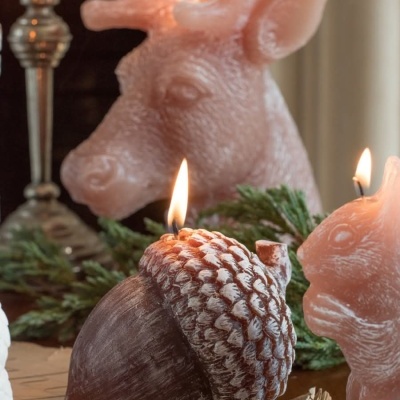 Velas decorativas em formas de cabeça de veado, coelho e bolota acesas sobre mesa com raminhos verdes