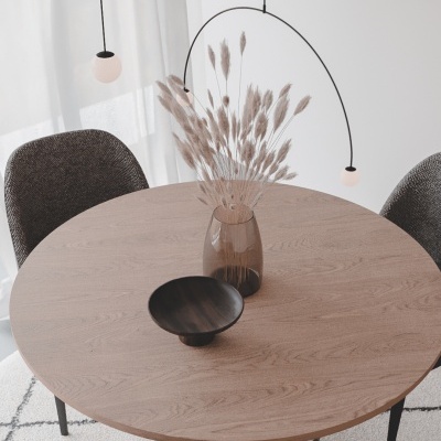 Mesa de madeira redonda com vaso e flores secas, rodeada por cadeiras cinzentas com cortinas brancas ao fundo