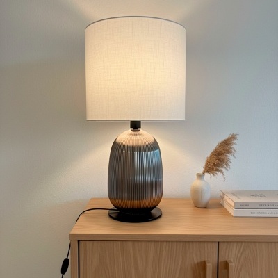 Abajur com base de vidro estriado acinzentado e cúpula branca sobre móvel de madeira clara com vaso e livros