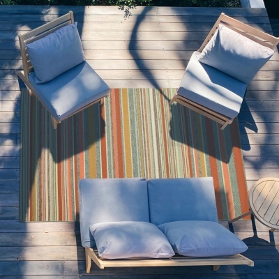 Móveis de jardim em madeira clara com almofadas cinza claro, tapete com riscas coloridas em deck de madeira