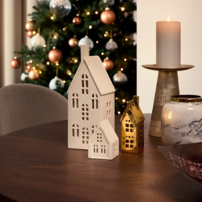 Casas decorativas em cerâmica branca e dourada numa mesa de madeira com vela acesa e árvore de Natal ao fundo