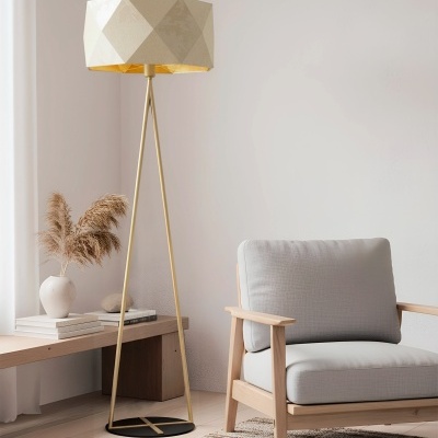 Lâmpada de pé dourada com abajur bege geométrico ao lado de poltrona cinza e mesa com vaso e livros.