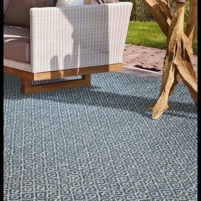 Sofá de exterior com braços em ráfia branca e almofadas cinzentas sobre tapete azul com padrão geométrico em ambiente exterior