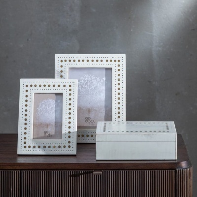 Dois porta-retratos e uma caixa brancos com detalhes dourados sobre móvel de madeira escura