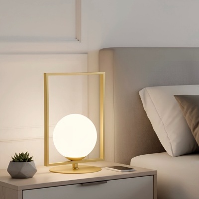 Abajur metálico dourado com esfera luminosa branca numa mesa de cabeceira junto a cama e planta suculenta.