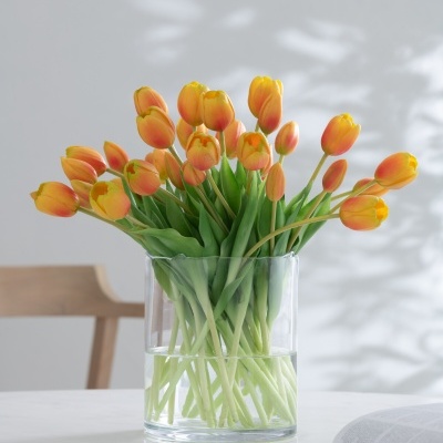 Vaso de vidro com tulipas amarelas e alaranjadas sobre mesa branca, cadeiras e livros ao fundo