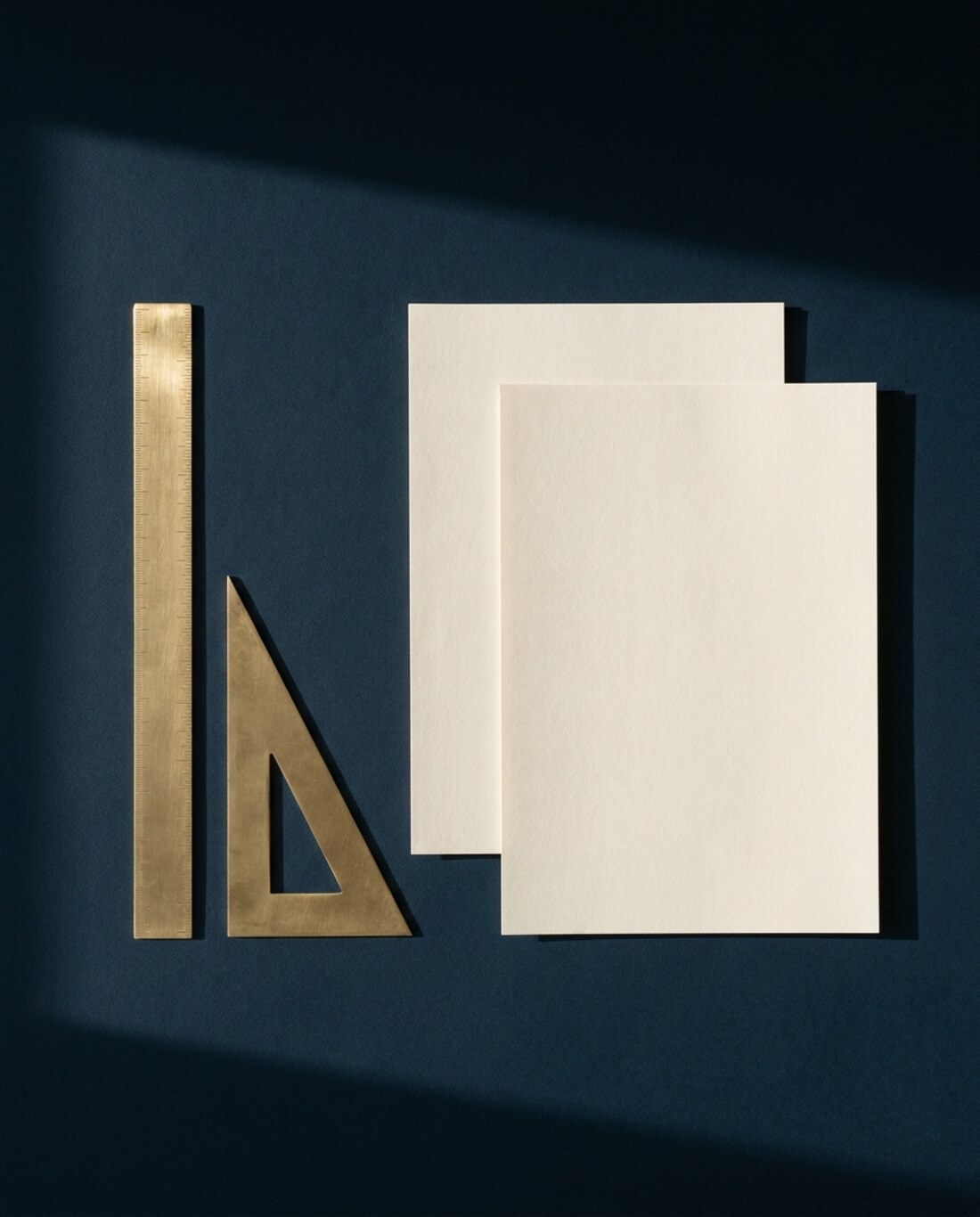 Régua e esquadro de latão sobre uma mesa azul-marinho, ao lado de duas folhas em branco