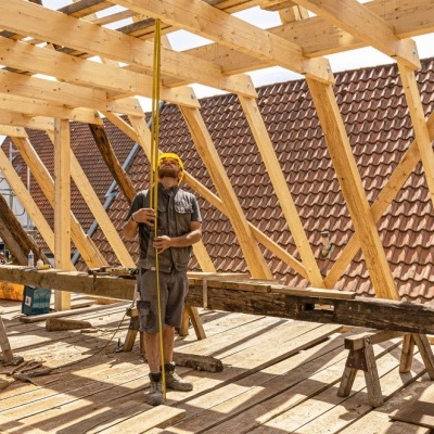 Estrutura de madeira em construção com trabalhador e telhados vermelhos ao fundo