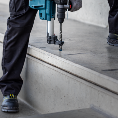 Furadeira elétrica azul a perfurar betão, operada por uma pessoa com calças pretas e sapatos de segurança.