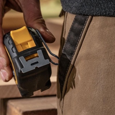 Mão a segurar fita métrica preta e amarela junto a calças castanhas