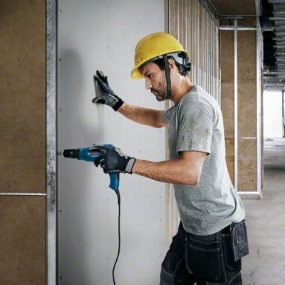Homem com capacete amarelo usando furadeira elétrica azul numa parede branca