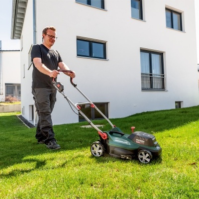 Homem a cortar a relva com corta-relvas elétrico verde em jardim em frente a casa branca