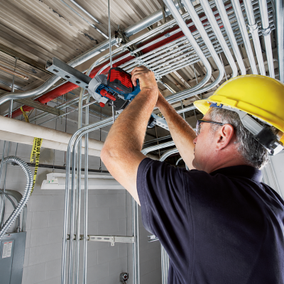Homem com capacete amarelo usando ferramenta elétrica em estrutura metálica no teto de sala industrial